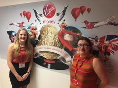 DB standing to left, and Vick from Virgin Money Bank standing to the right. Between them on the wall is a big mural photo collage with lots of Virgin services and products with their motto 'On a quest to make banking better'
