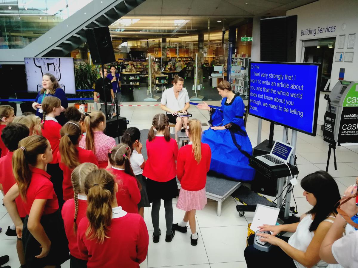 A group of children near the stage watching PAD productions actress dressed up as Harriet Martineau, Deaf Author and her assistant to her left. There is also a lipspeaker on the left side of the stage and a speech to text reporter sitting down on the right side which is projected on a screen what is being said